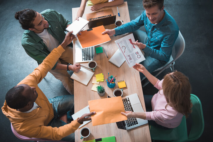 Creative teamwork. Top view of excited young workers are transferring documents to each other while sitting at table. They are expressing gladness while working together